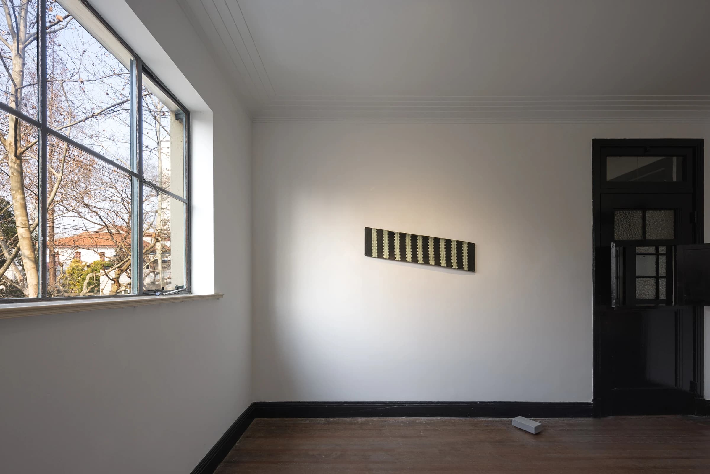 A horizontal painting on a white gallery wall beside a window, with natural light across the artwork and wooden floor.